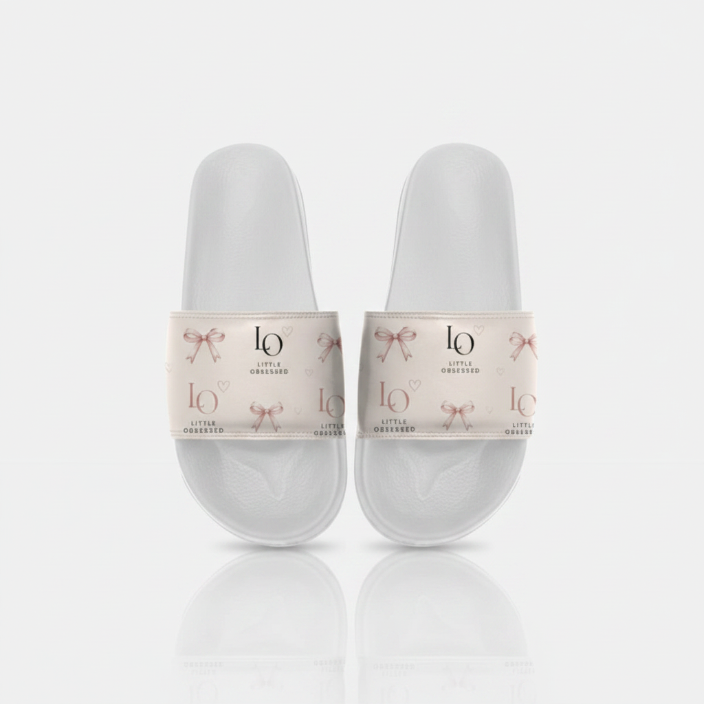 White slide sandals with blush-pink strap, rose-gold bows and Little Obsessed LO branding on a clean studio background