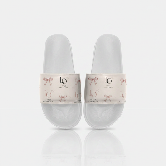 White slide sandals with blush-pink strap, rose-gold bows and Little Obsessed LO branding on a clean studio background